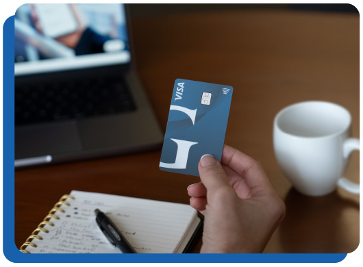 A member holding a Guardian Credit Union Visa Credit card while taking notes in a notepad with a laptop open in front and a coffee mug at the side.